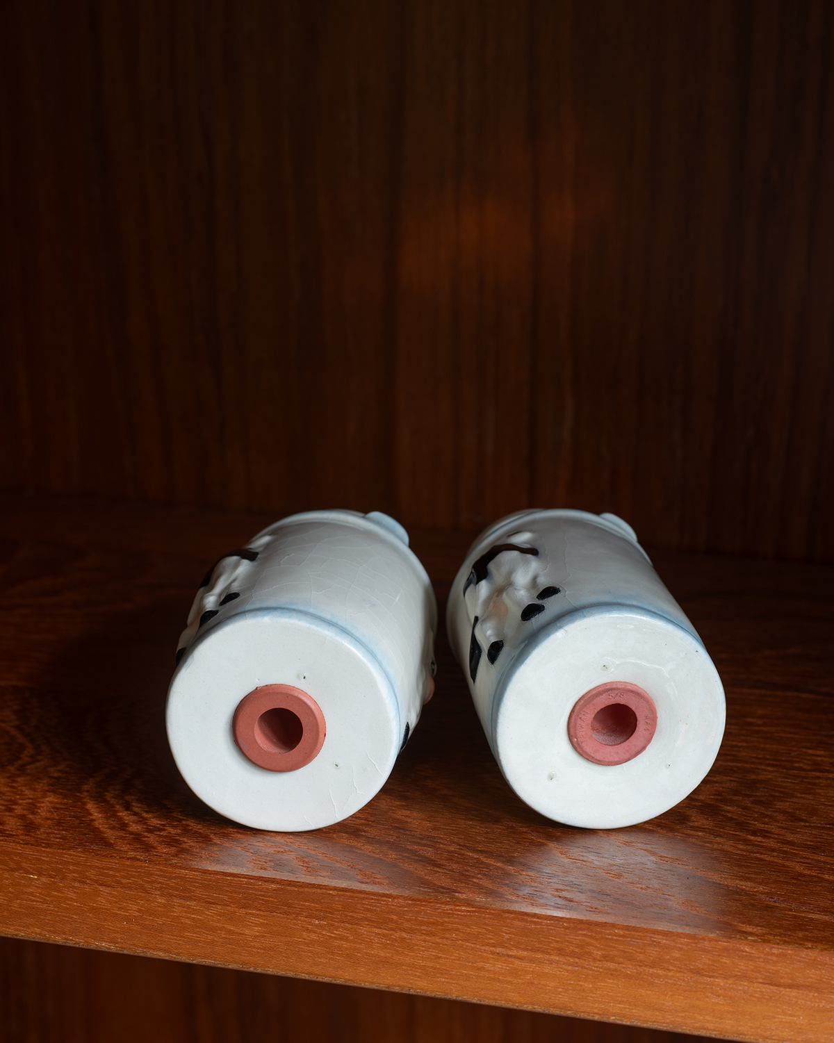 Two white ceramic cylindrical vases with black floral patterns and pink centers, lying on their sides on a wooden shelf against a dark wood background.