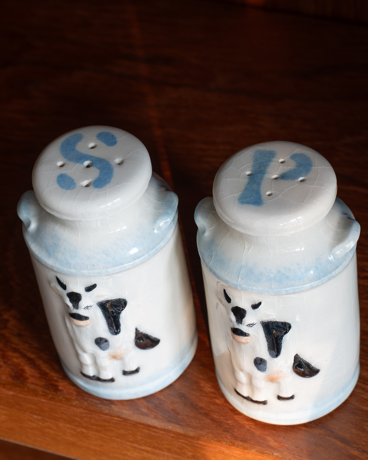 A pair of ceramic salt and pepper shakers with cow designs, sitting on a dark wooden surface. The shakers are white with light blue accents at the base and top, featuring black and white cow faces and blue letters 'S' and 'P' on the lids.