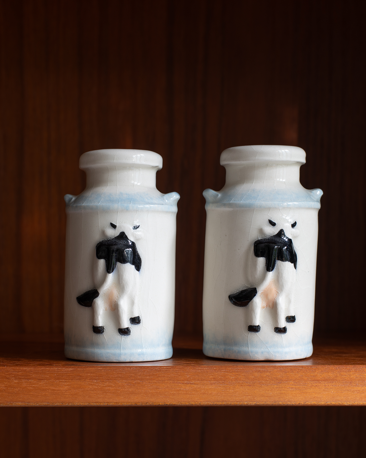 Two small white ceramic bottles with black and white cow figures and light blue bands near the top, displayed side by side on a wooden surface against a dark wood background.