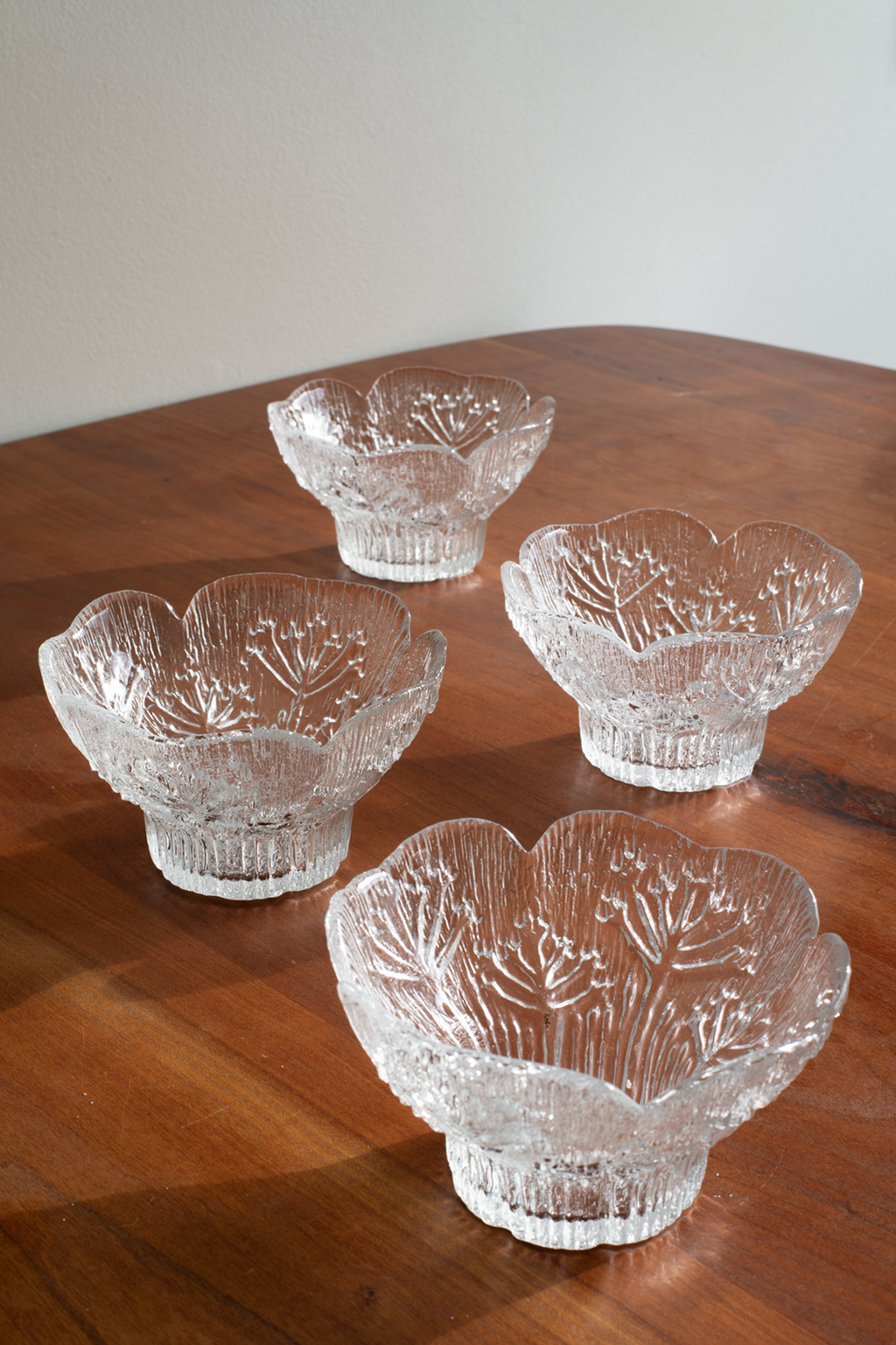 Four clear cut-glass bowl-like serving pieces with a floral or leaf-like pattern, arranged on a wooden tabletop against a plain light wall.