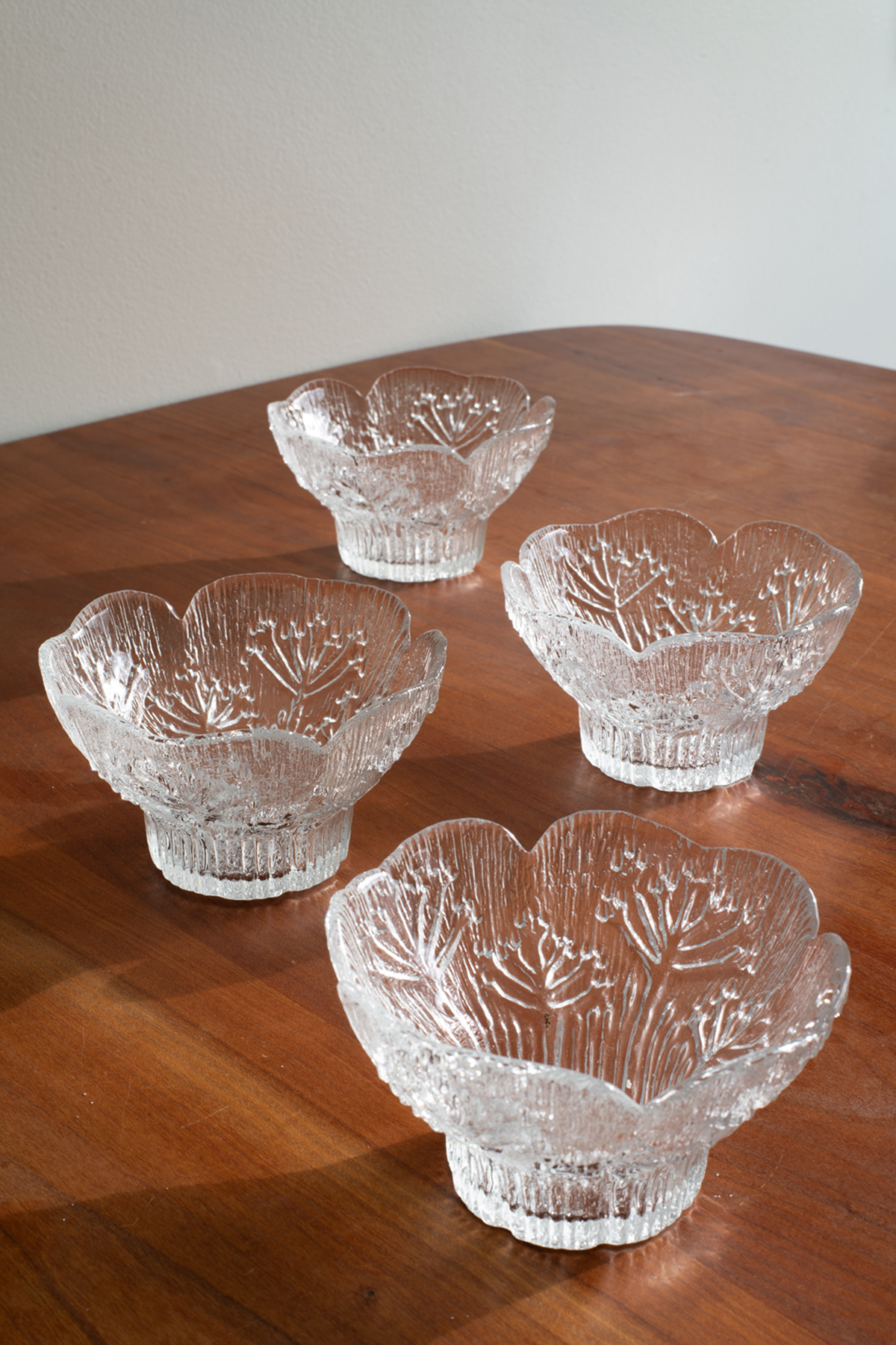 Four clear cut-glass bowl-like serving pieces with a floral or leaf-like pattern, arranged on a wooden tabletop against a plain light wall.