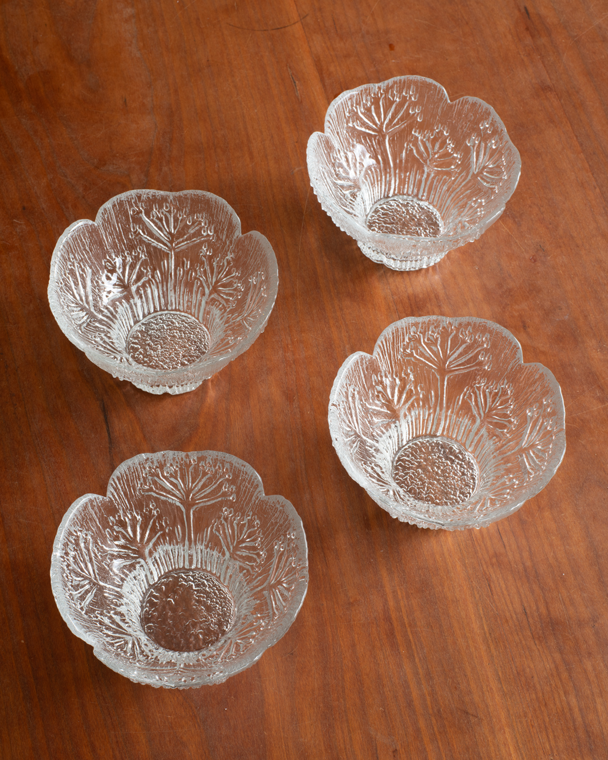 Four clear glass bowls with scalloped edges and etched floral patterns, arranged on a warm brown wooden surface.