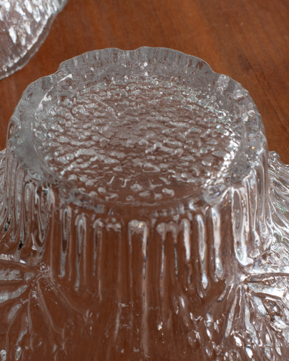 A close-up of a clear textured glass bowl or dish with a scalloped rim, photographed on a brown wooden surface.