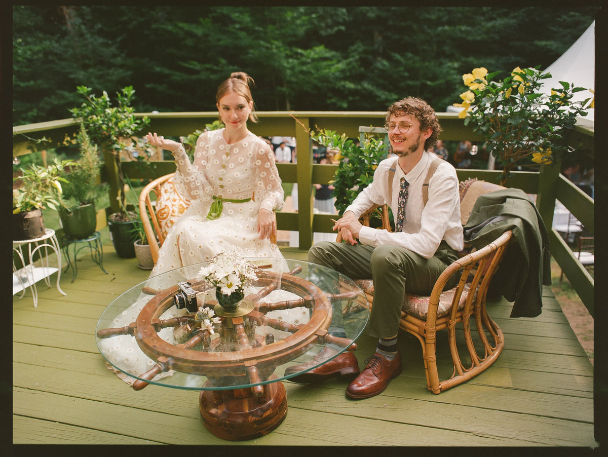 A bride in a white lace wedding dress and a groom in a white shirt and olive green trousers sit on wicker chairs on a wooden deck, sharing a glass table with a ship's wheel base. The background shows a lush green forest and garden plants.