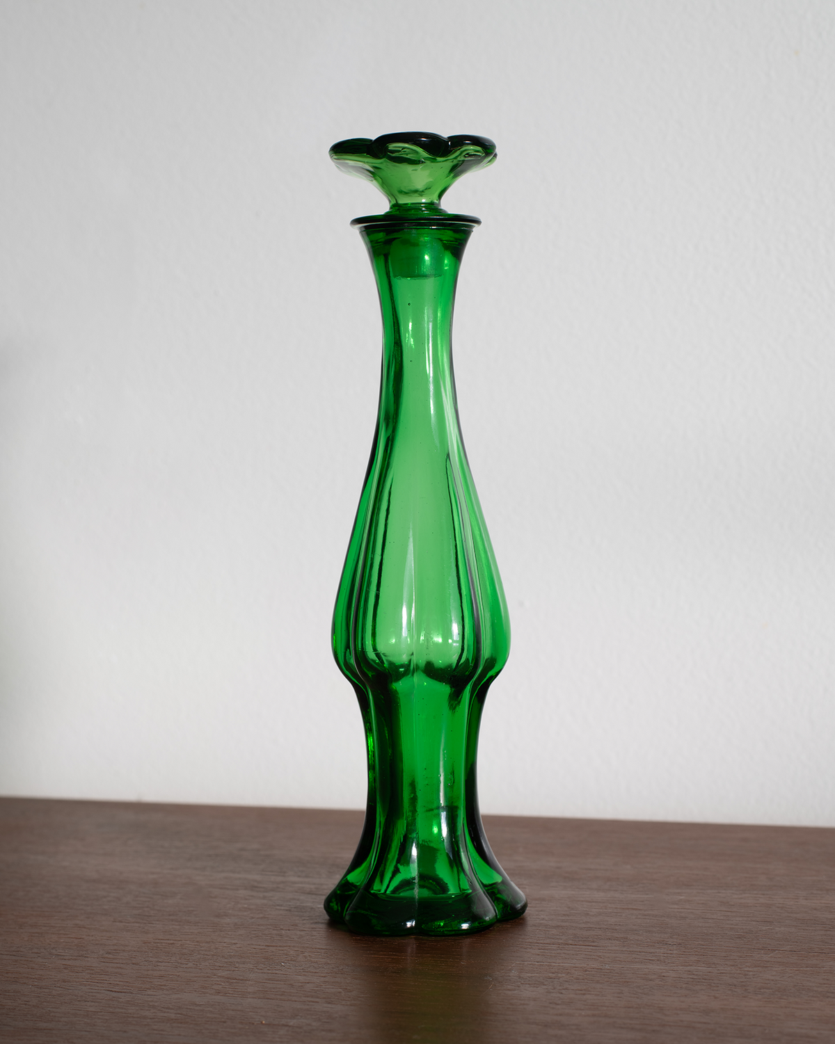 A tall, slender green glass vase with a flared flower-shaped top, standing on a wooden surface against a plain white wall.