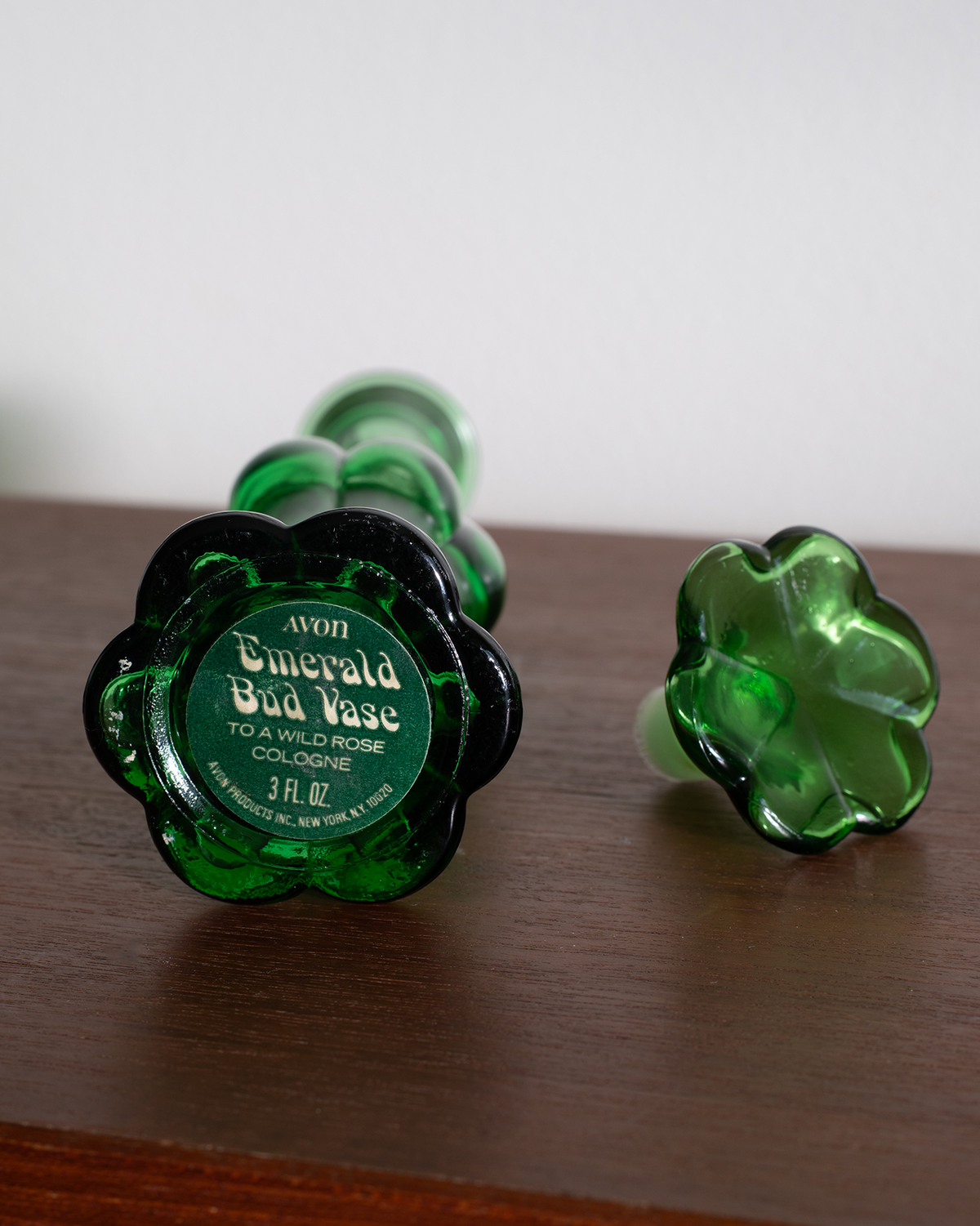 A green glass bud vase with a clover-shaped stopper, shown on a wooden surface against a light gray background. The bottom of the vase is visible and reads 'AYON Emerald Bud Vase'.
