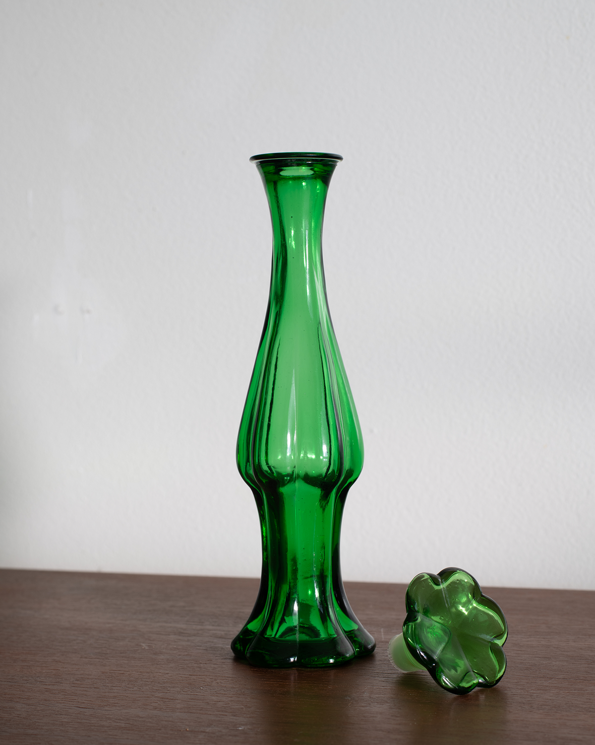 A tall, fluted green glass vase stands on a wooden surface against a plain white wall, with a matching green flower-shaped glass object resting beside it.