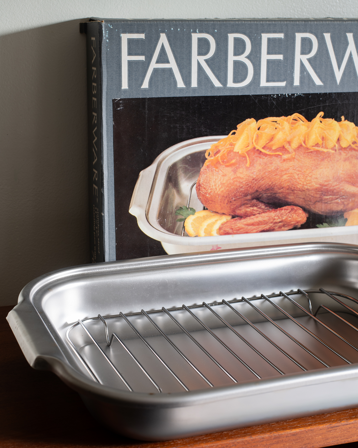 A silver metal roasting pan with a wire rack sits on a wooden surface in front of a Farberware cookbook, with a baked bird visible on the book cover.
