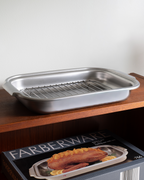 A stainless steel baking pan with an integrated wire rack, shown resting on a wooden shelf above its Farberware product box.