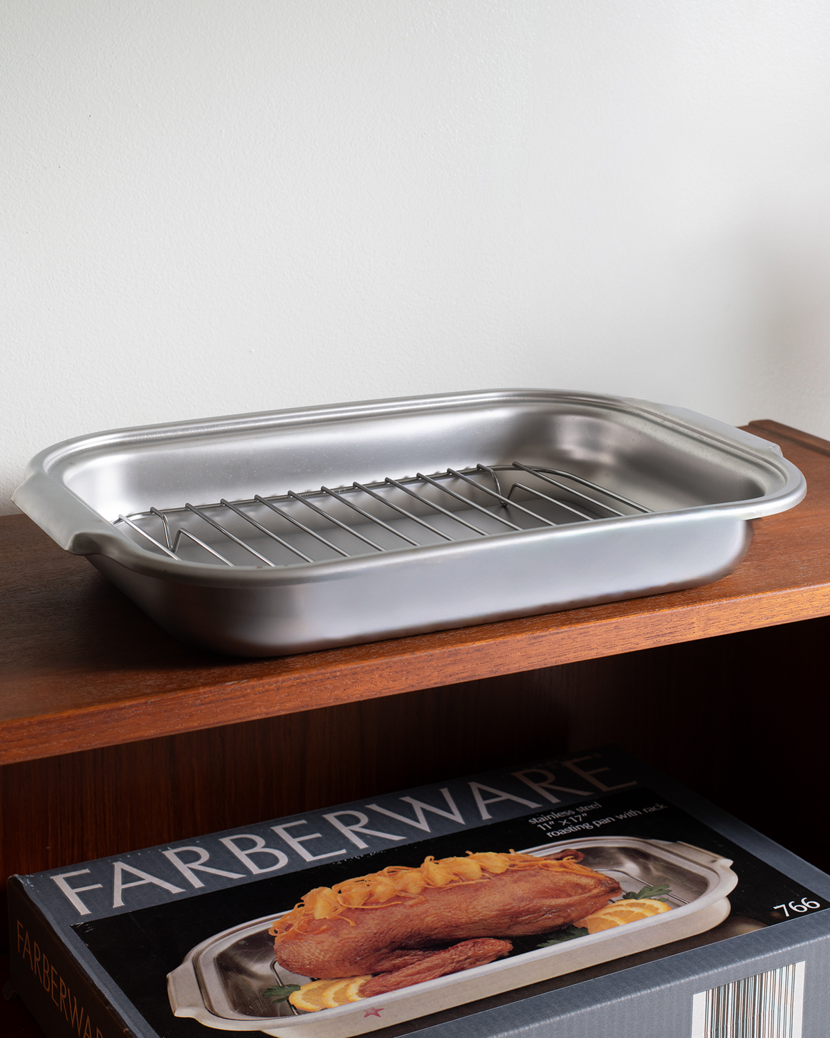 A stainless steel baking pan with an integrated wire rack, shown resting on a wooden shelf above its Farberware product box.