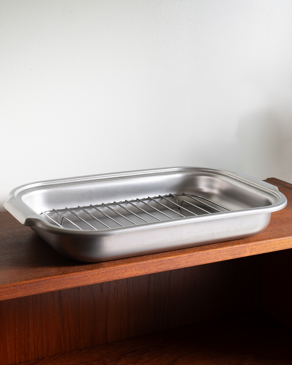A stainless steel food pan with an integrated wire cooling rack, resting on a wooden shelf against a plain white wall.
