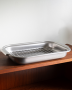 A stainless steel food pan with an integrated wire cooling rack, resting on a wooden shelf against a plain white wall.