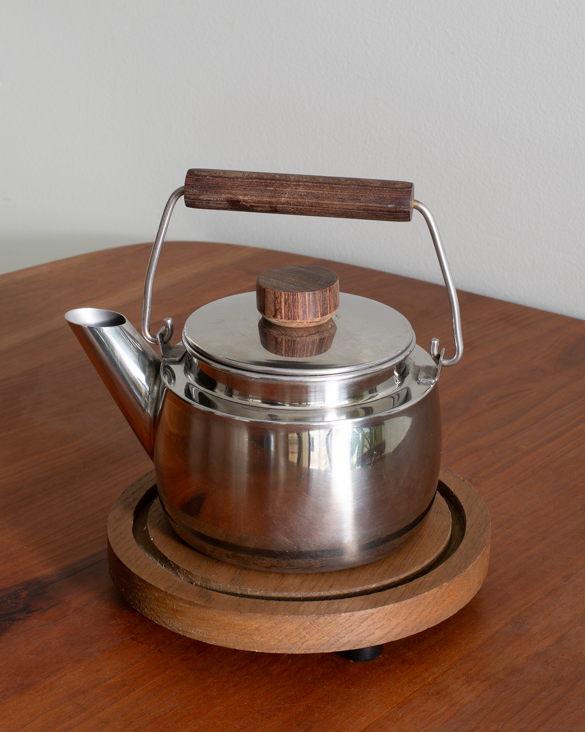 A stainless steel teapot with a dark brown wooden handle and lid knob, resting on a round wooden trivet on a wooden table against a plain light gray wall.