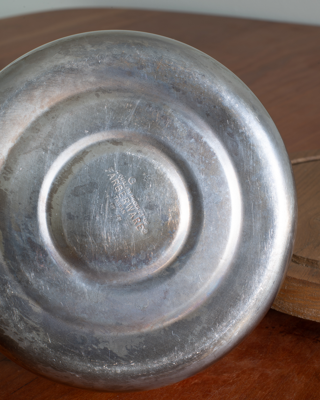 A close-up of the bottom of a round silver metal plate or shallow pan, showing a stamped maker's mark in the center, resting on a wooden surface.