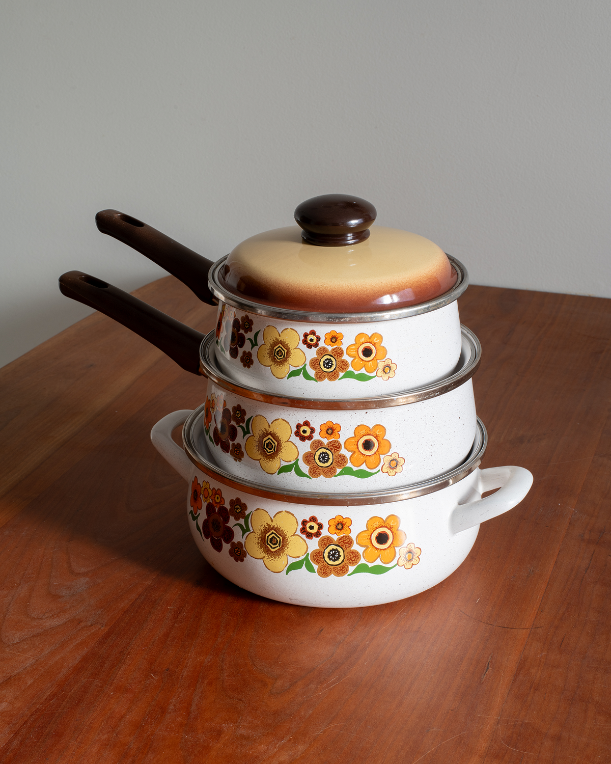 A stack of three white enamel nesting cookware pots with lids, decorated with a retro yellow, orange, and brown floral pattern, resting on a wooden table against a neutral gray background.