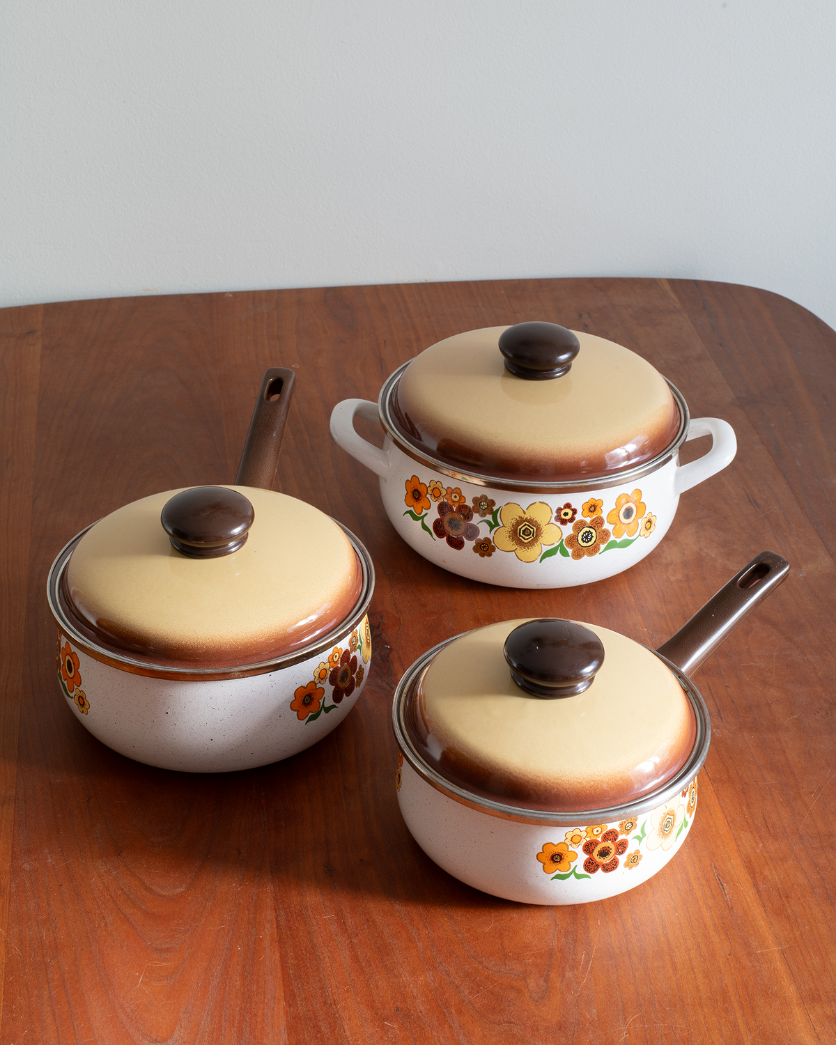 Three vintage enamel cookware pots with matching lids, featuring a white base, a warm beige lid, and a colorful floral pattern, arranged on a wooden table.