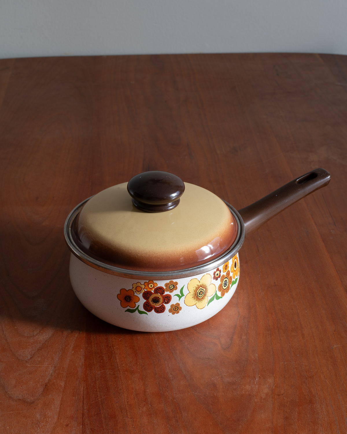A vintage-style enamel saucepan with a lid, featuring a white body, brown handle and lid knob, and a colorful floral pattern around the lower rim, set on a wooden table.