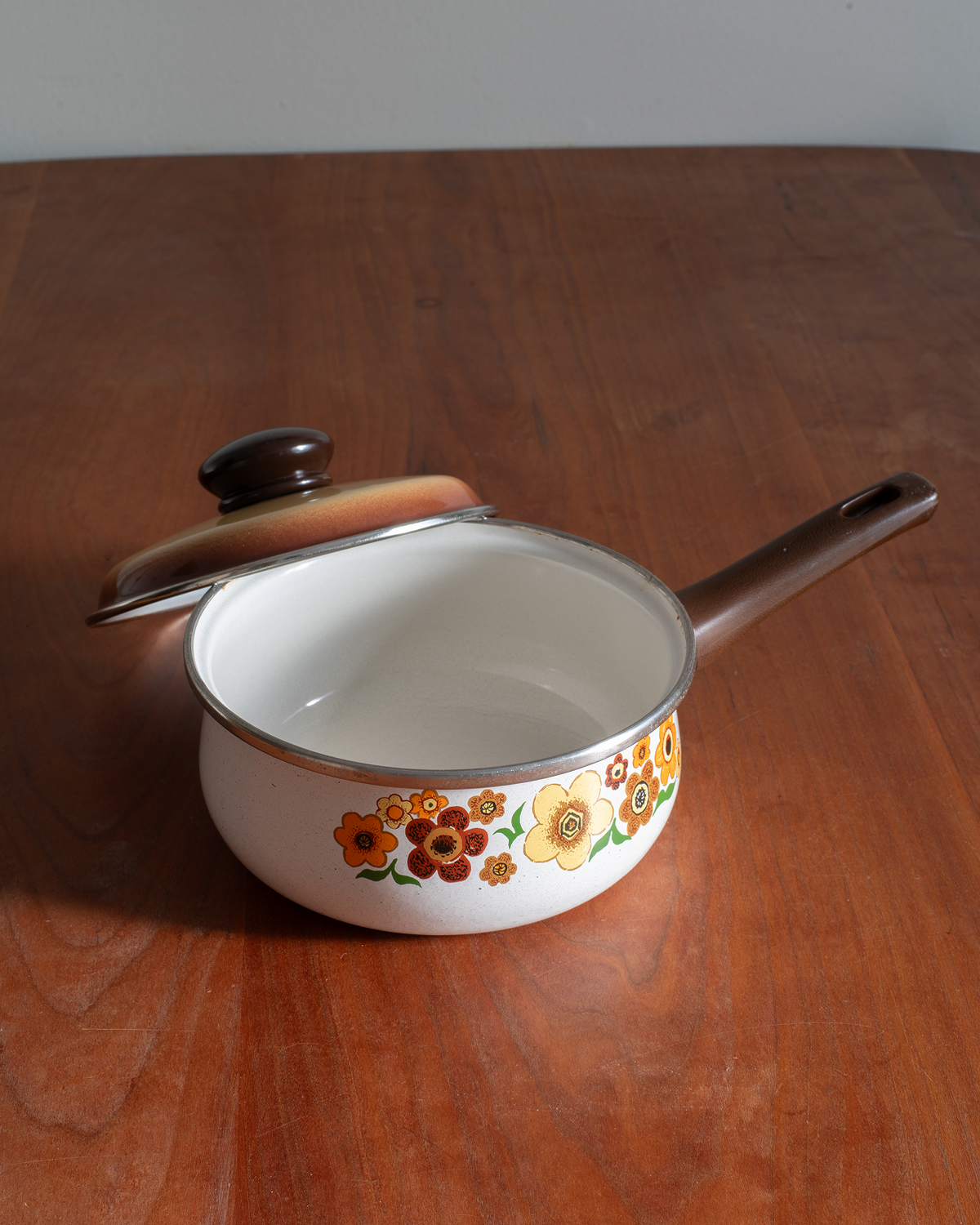 A vintage white enamel saucepan with a brown handle and lid, decorated with a colorful floral pattern around the lower half, resting on a wooden table.