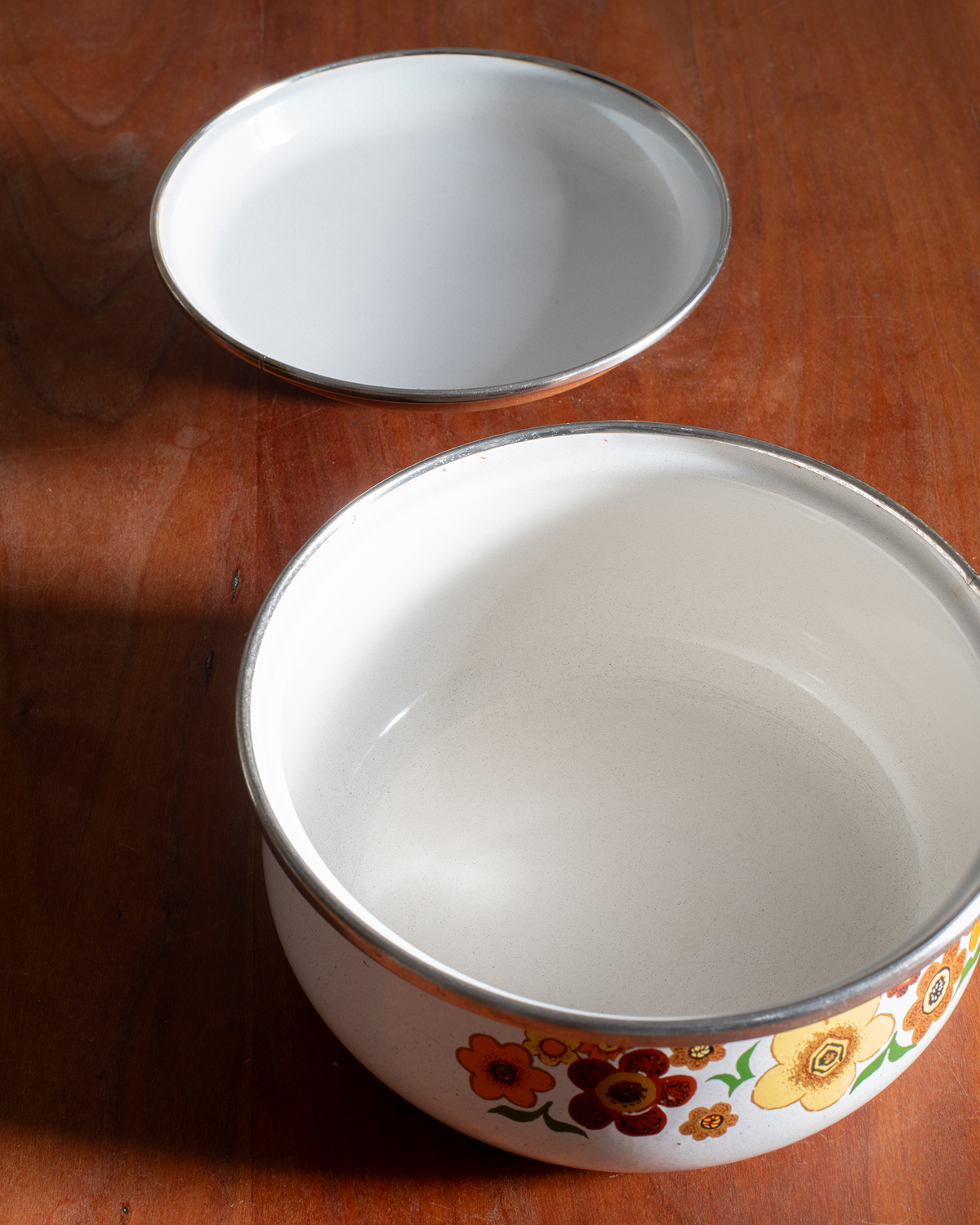 A white enamel bowl with a silver rim and a colorful floral pattern around the base, shown alongside its matching lid on a wooden tabletop.