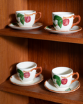 Four white ceramic teacups with saucers, decorated with a hand-painted red apple motif with green leaves and brown handles, displayed on a wooden shelf.