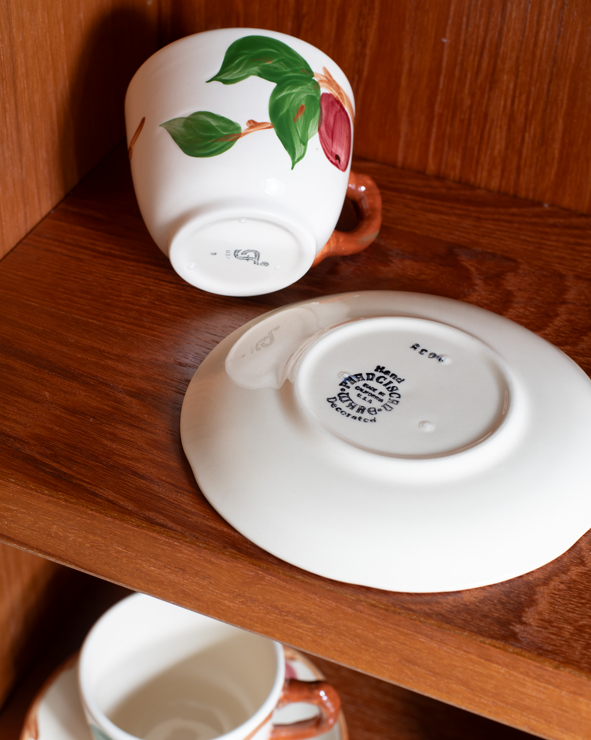 A white ceramic teacup with a green leaf and red fruit illustration sits upside down on a wooden shelf next to its matching saucer, with another cup partially visible below.