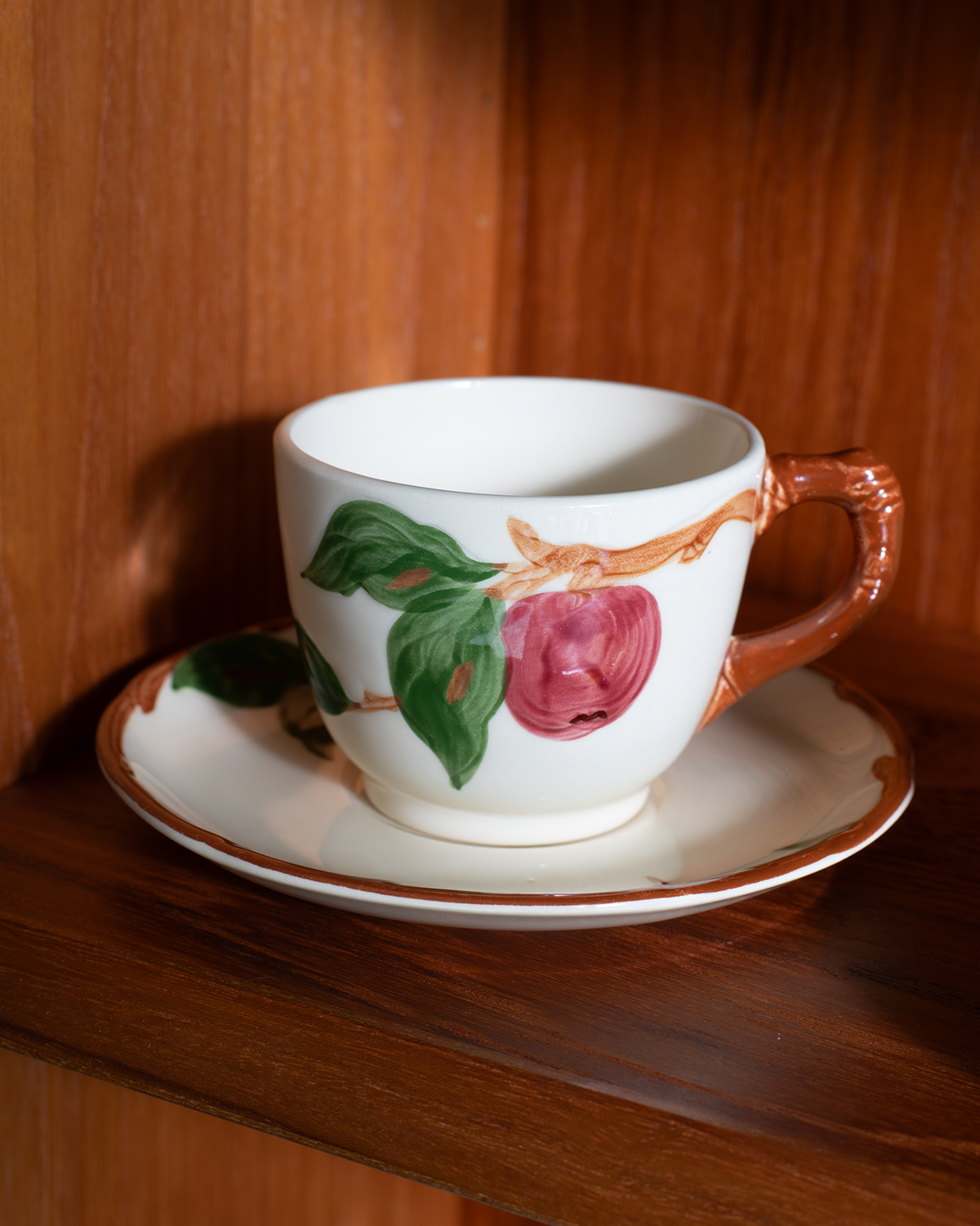 A white ceramic teacup and saucer set with a hand-painted apple design featuring green leaves and a pinkish-red apple, displayed on a wooden shelf.