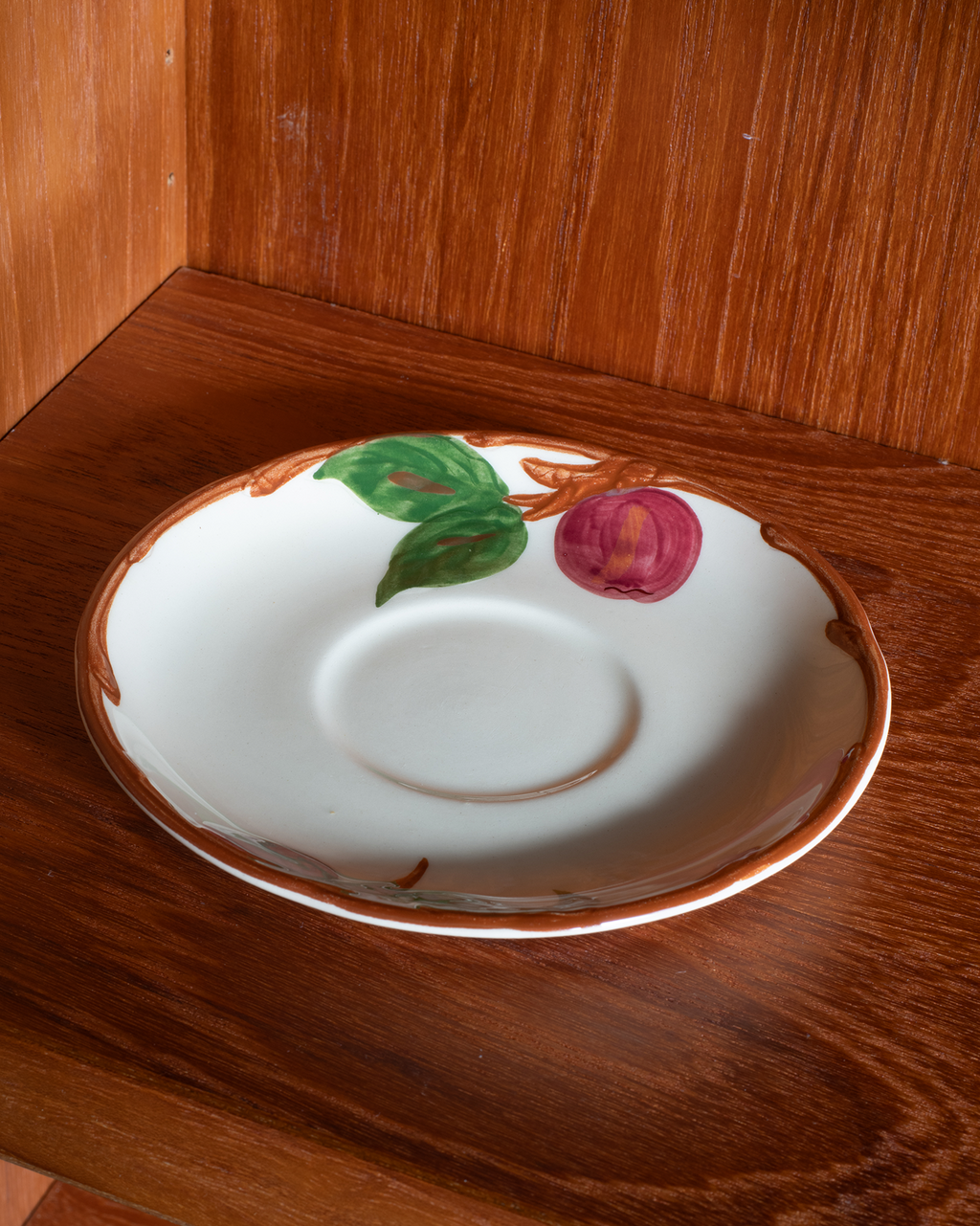 A white ceramic saucer with a hand-painted green leaf, a pinkish-purple flower, and a brown rim, displayed on a wooden shelf.