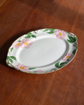 An oval white ceramic serving platter with a scalloped rim, decorated with pink flowers and green leaves, placed on a dark brown wooden table.