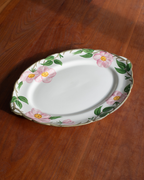 An oval white ceramic serving platter with a scalloped rim, decorated with pink flowers and green leaves, placed on a dark brown wooden table.