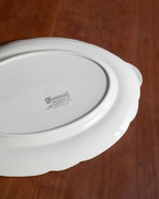 A white ceramic serving platter with a scalloped rim, photographed from above on a dark brown wooden surface. The brand name 'Frondsong' is printed in the center.