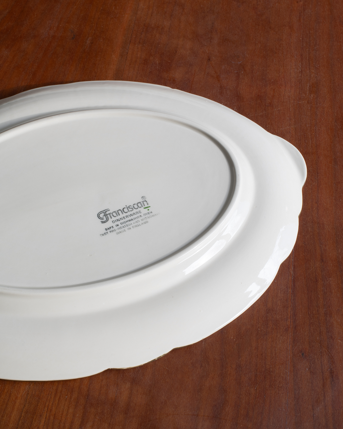 A white ceramic serving platter with a scalloped rim, photographed from above on a dark brown wooden surface. The brand name 'Frondsong' is printed in the center.