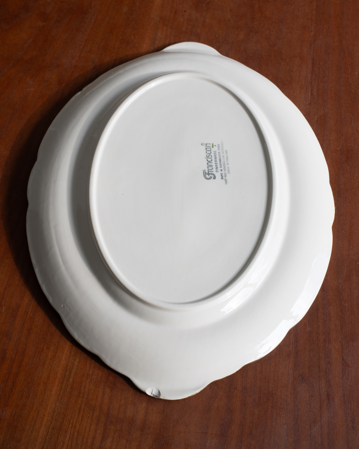 An oval white ceramic serving platter with a scalloped rim, decorated with pink flowers and green leaves, placed on a dark brown wooden table.