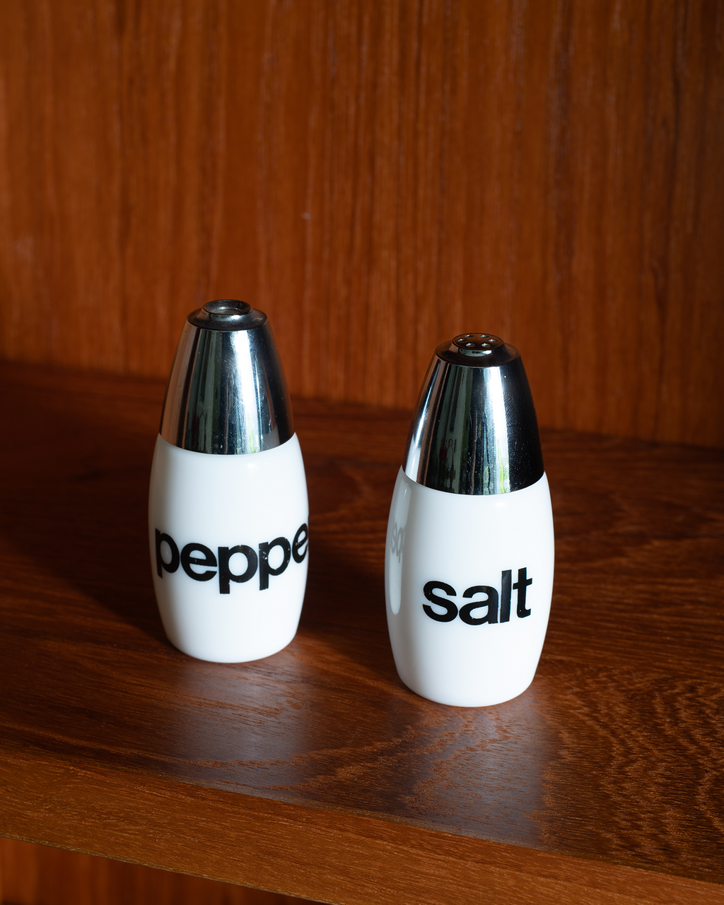 Two white ceramic pepper and salt shakers with shiny silver metal tops, standing on a wooden surface against a wooden background. The pepper shaker is labeled 'peppe' and the salt shaker is labeled 'salt'.