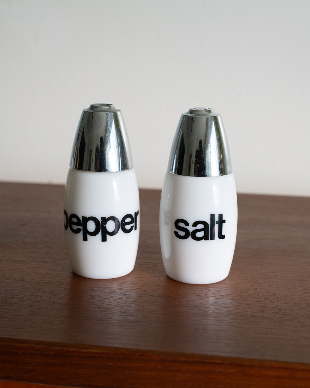 Two white ceramic pepper and salt shakers with shiny silver metallic caps, standing on a dark wooden surface against a plain light background.