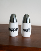 Two white ceramic pepper and salt shakers with shiny silver metallic caps, standing on a dark wooden surface against a plain light background.