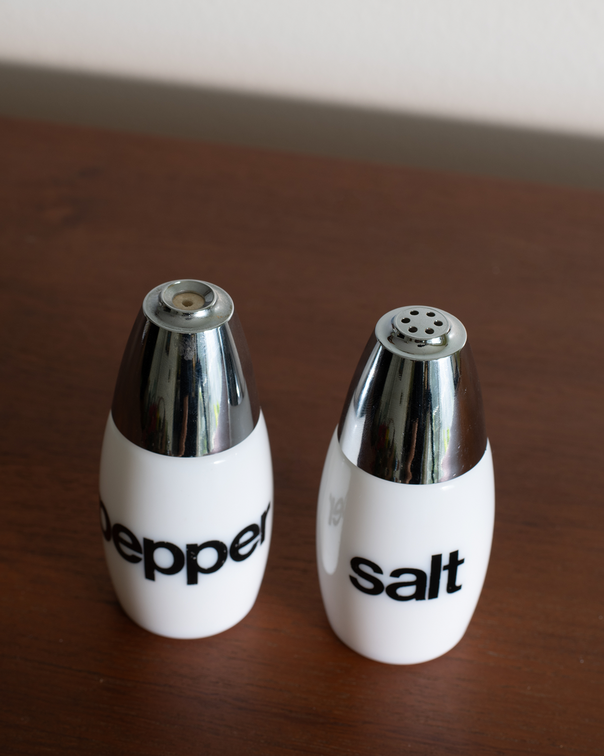 A pair of white ceramic pepper and salt shakers with shiny chrome caps, standing on a dark wooden tabletop.