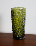A translucent olive green glass tumbler with a raised bubble pattern, standing on a dark wooden surface against a plain light gray wall.