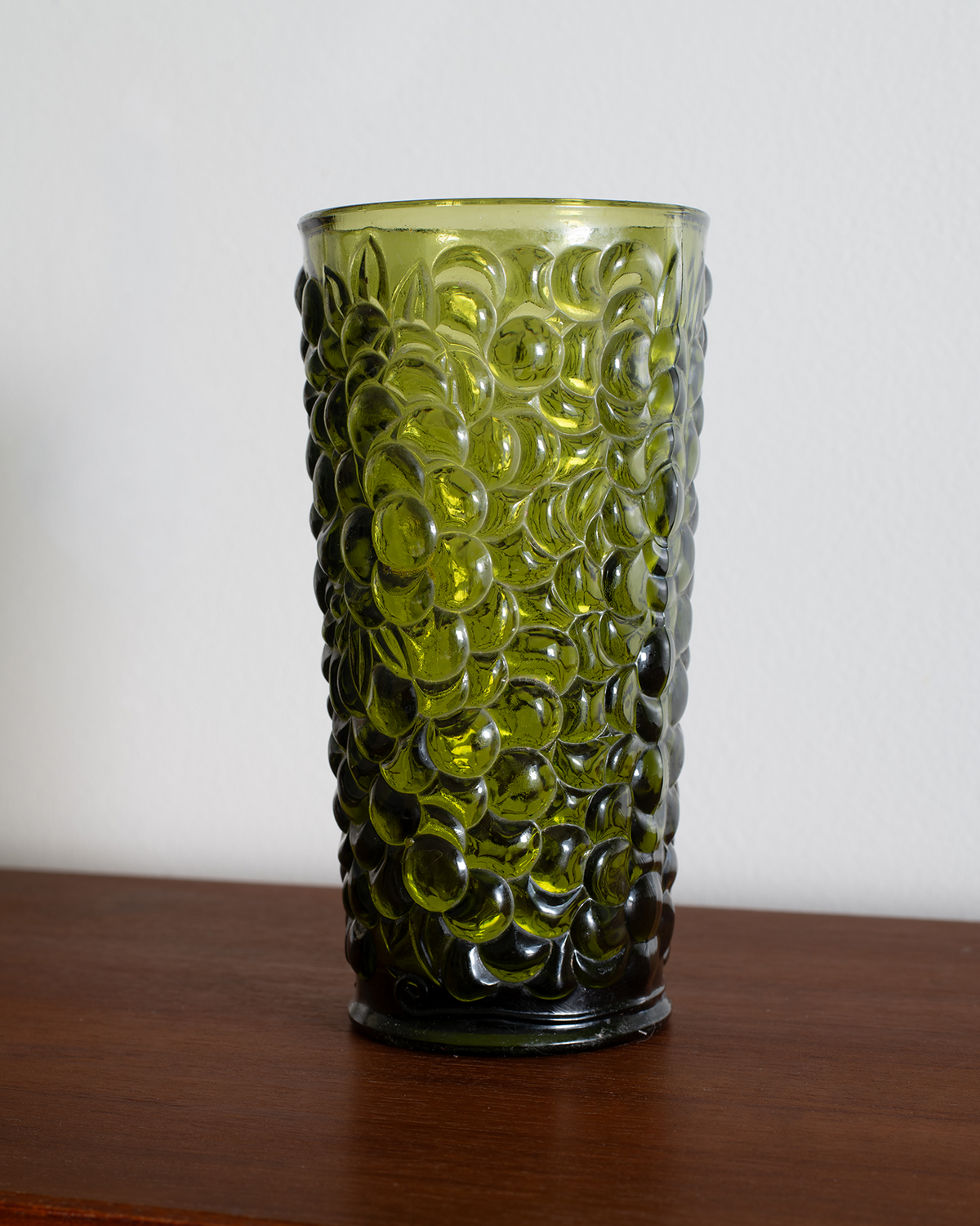 A translucent olive green glass tumbler with a raised bubble pattern, standing on a dark wooden surface against a plain light gray wall.