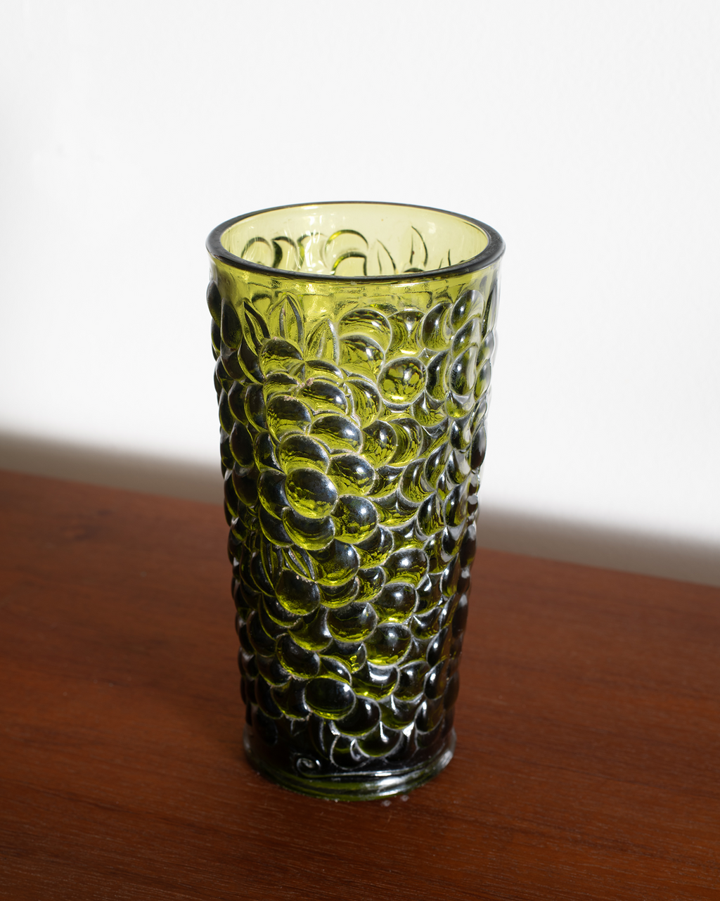 A textured green glass drinking cup with a bubble-like embossed pattern, standing on a wooden surface against a plain white background.