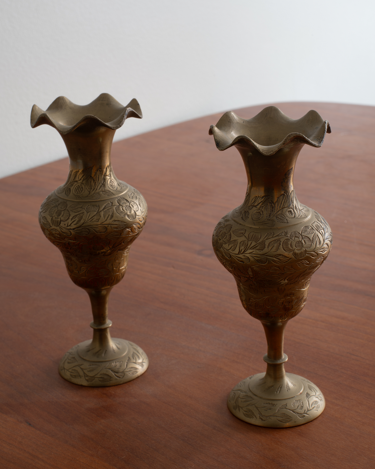 Two ornate brass pedestal vases with flared, ruffled rims and embossed floral patterns, displayed on a wooden table against a plain light background.