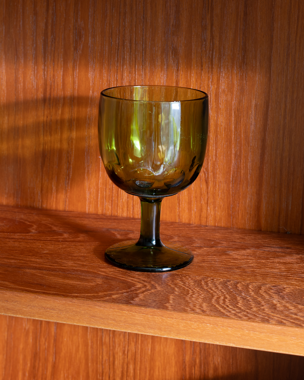 An olive green tinted glass goblet with a wide bowl and a short stem, resting on a wooden shelf against a matching wood-paneled background.