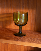 An olive green tinted glass goblet with a wide bowl and a short stem, resting on a wooden shelf against a matching wood-paneled background.