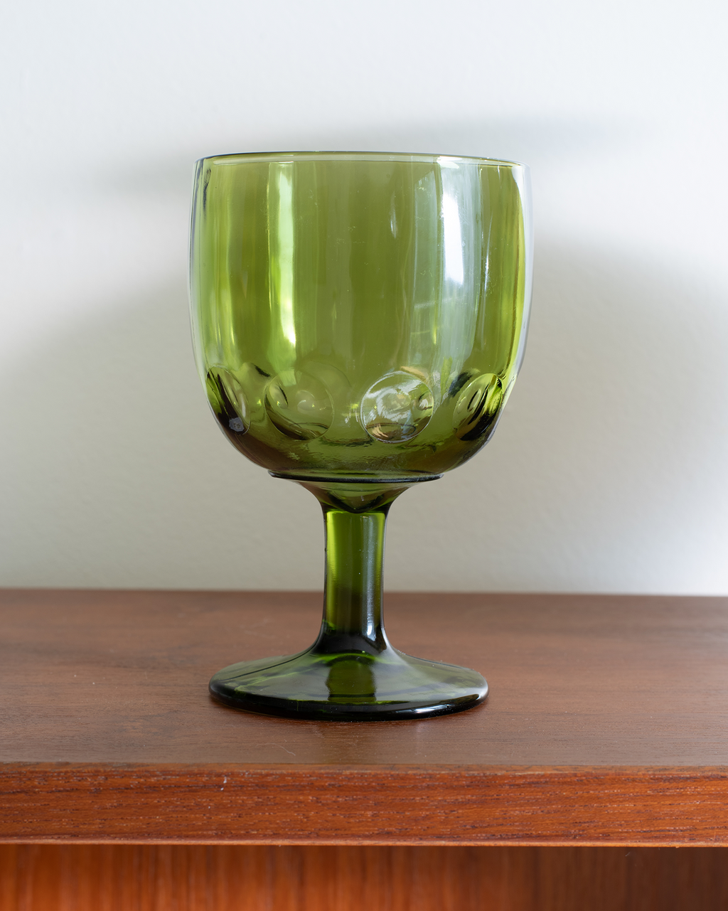 An olive green glass goblet with a wide bowl, textured base, and stem, resting on a wooden surface against a plain light gray wall.