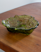 An olive green leaf-shaped glass bowl with embossed leaf patterns, resting on a wooden tabletop.