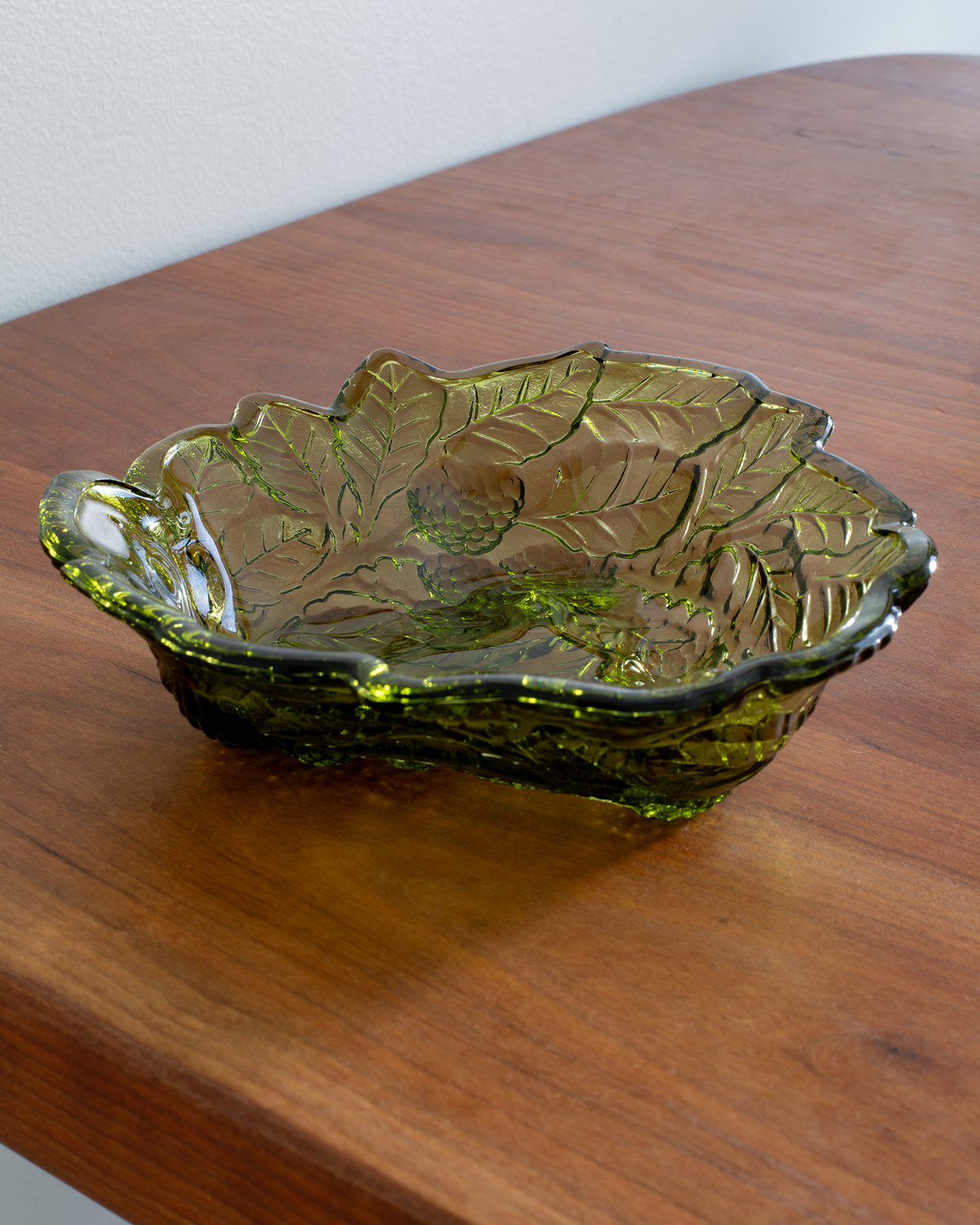 An olive green leaf-shaped glass bowl with embossed leaf patterns, resting on a wooden tabletop.