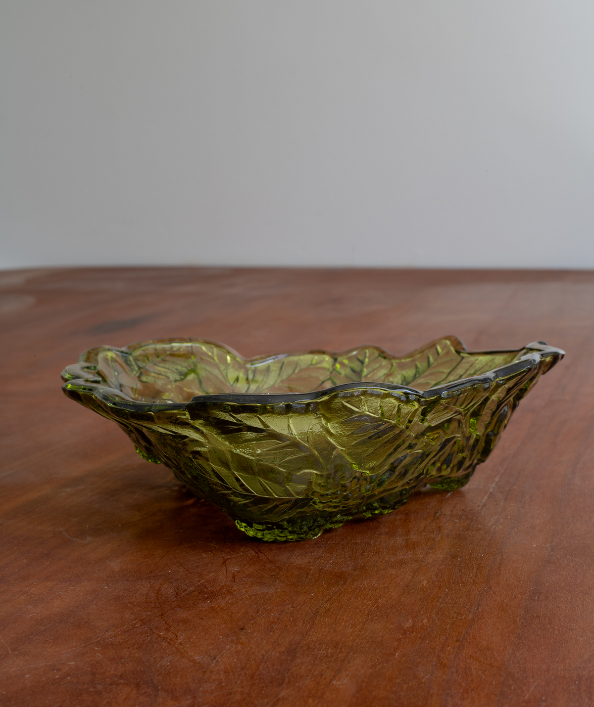 An olive green textured glass bowl with a scalloped rim and footed base, set on a wooden table against a plain light gray background.