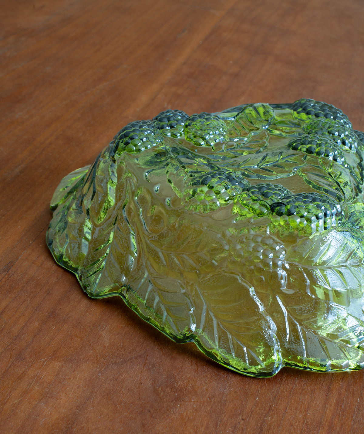 A vintage green glass bowl with an embossed grapevine pattern, resting on a wooden tabletop.