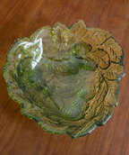 A scalloped, leaf-patterned glass bowl with a green and amber-brown tint, photographed from above on a wooden tabletop.
