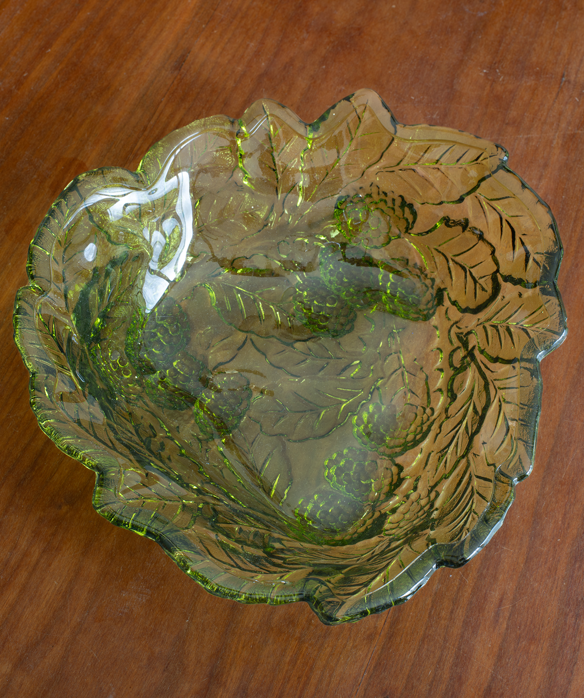 A scalloped, leaf-patterned glass bowl with a green and amber-brown tint, photographed from above on a wooden tabletop.