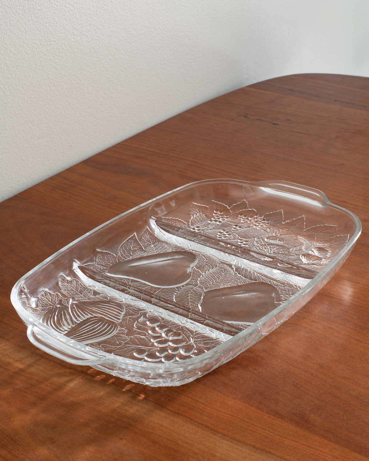 A clear glass divided serving tray with embossed floral and leaf patterns, resting on a wooden table against a light wall.